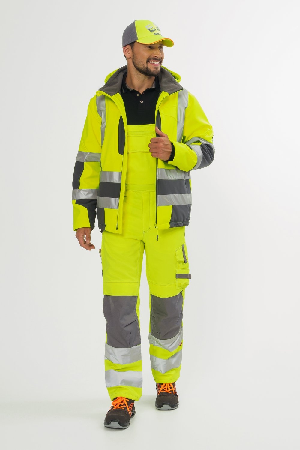 Mann trägt gelbe Arbeitsjacke mit Reflexstreifen, passende Latzhose mit grauen Einsätzen, schwarzes Poloshirt und Sicherheitsschuhe.