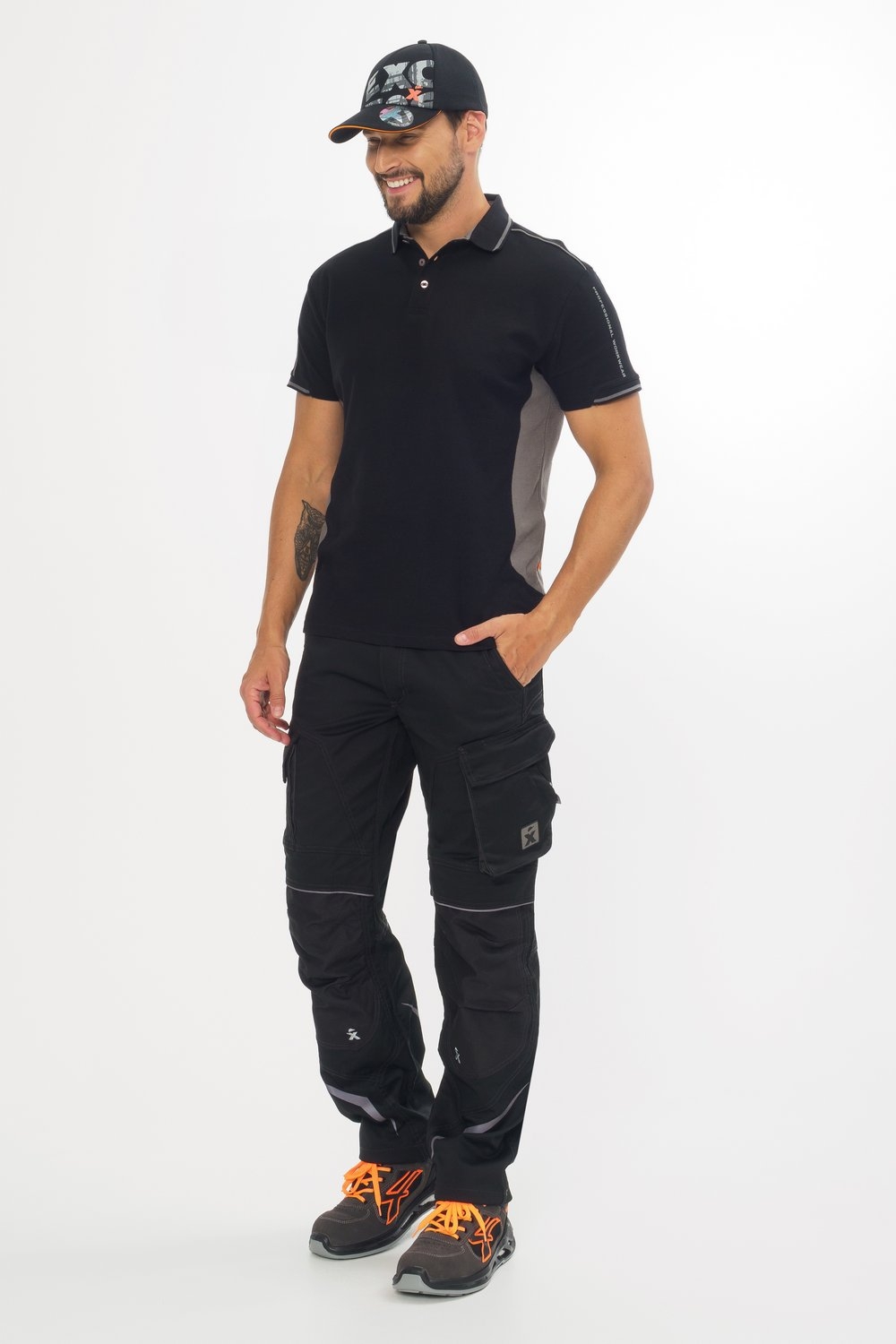 Mann trägt schwarzes Poloshirt, schwarze Arbeitshose mit Taschen, schwarze Cap und Sicherheitsschuhe mit orangefarbenen Details.