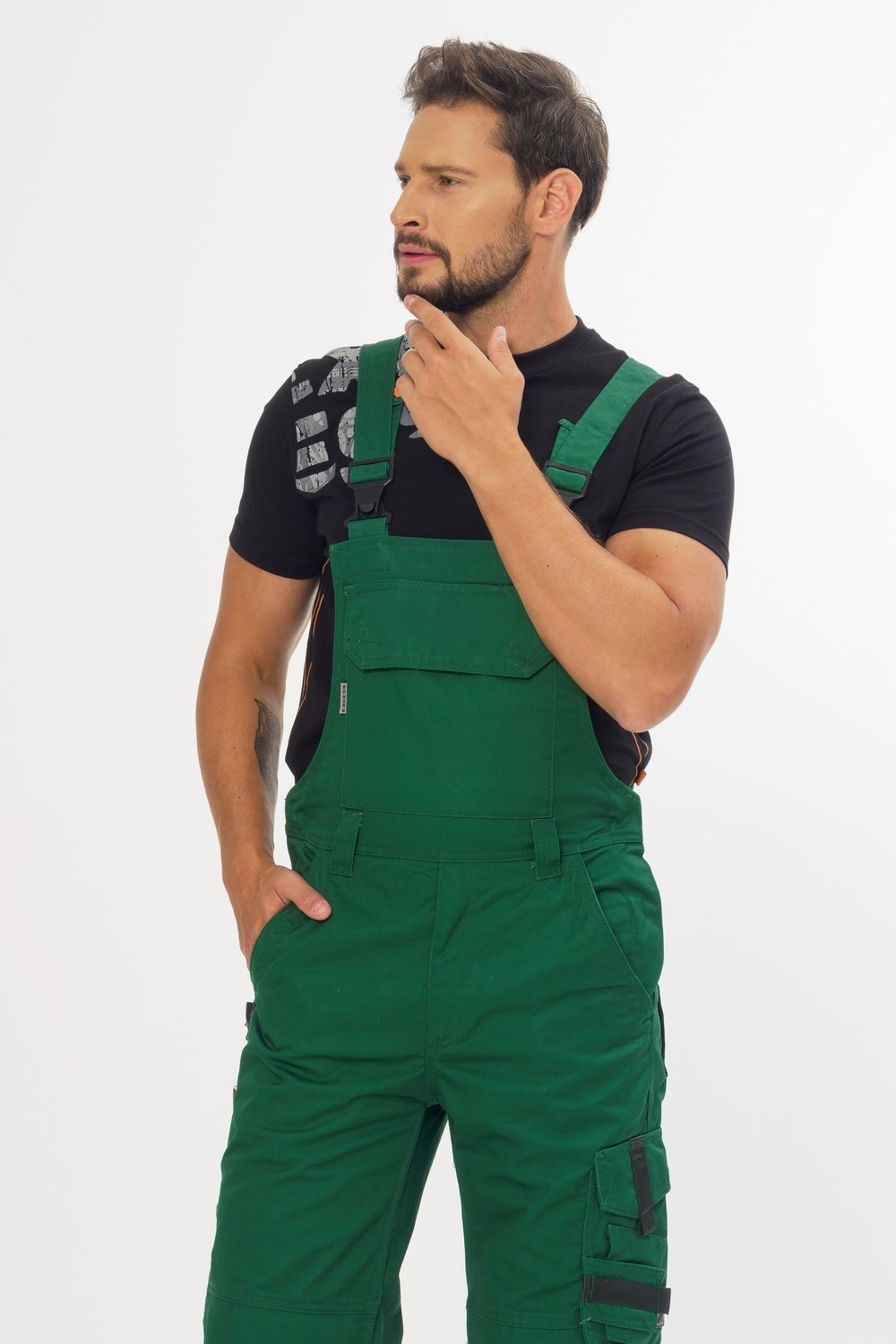 Handwerker in grüner Latzhose mit großer Brusttasche, seitlichen Cargotaschen und verstellbaren Trägern. Dazu ein schwarzes T-Shirt, geeignet für Arbeiten im Handwerk, Bau und Gartenbereich.