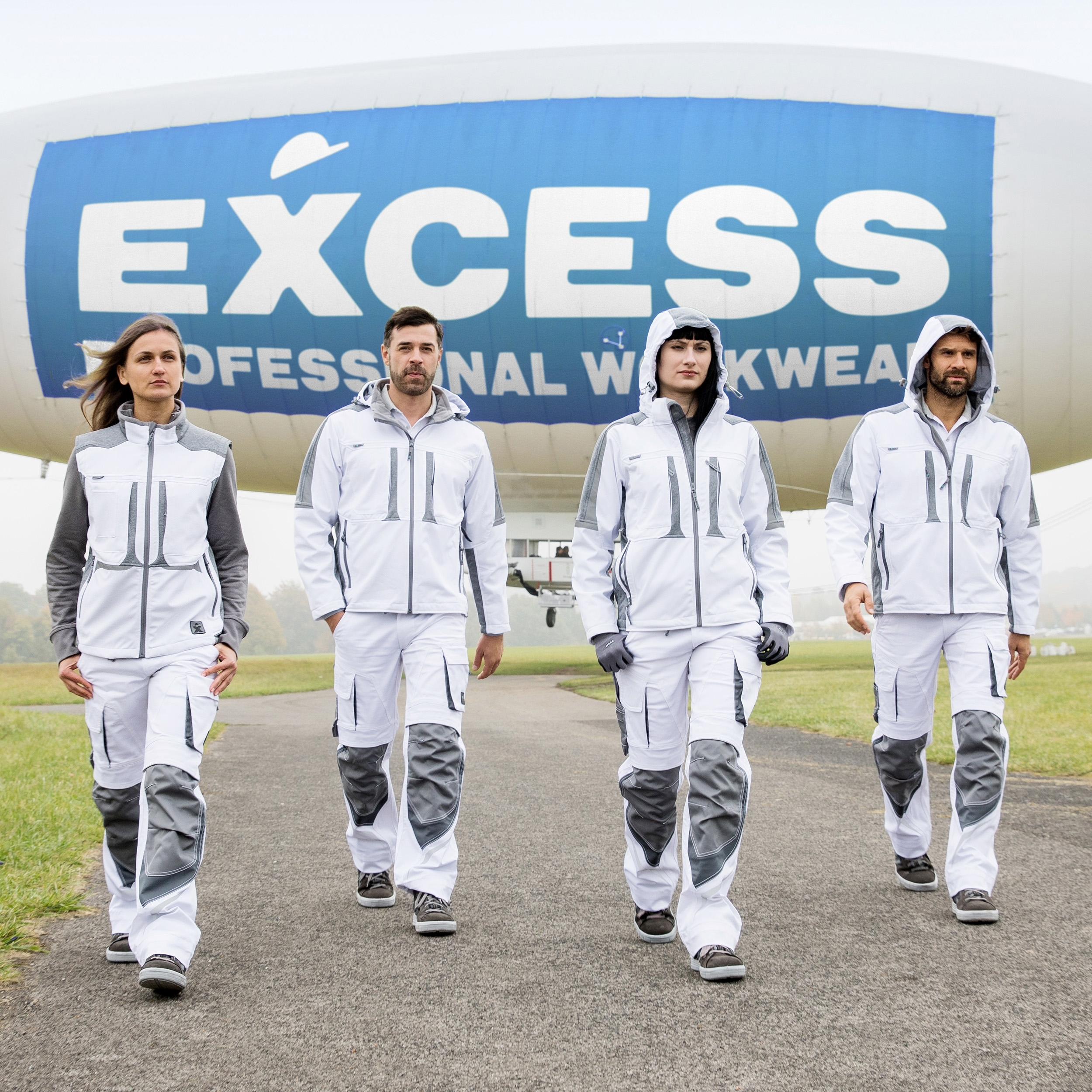 Vier Personen in weiß-grauer Arbeitskleidung laufen auf einer Straße, im Hintergrund ein EXCESS Workwear Luftschiff.