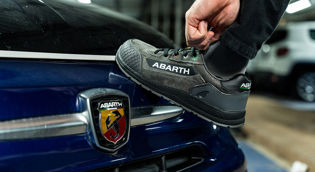 Ein grauer Abarth-Arbeitsschuh steht auf dem Heck eines blauen Autos mit dem Abarth-Logo. Eine Hand hält den Schuh leicht an der Ferse fest.