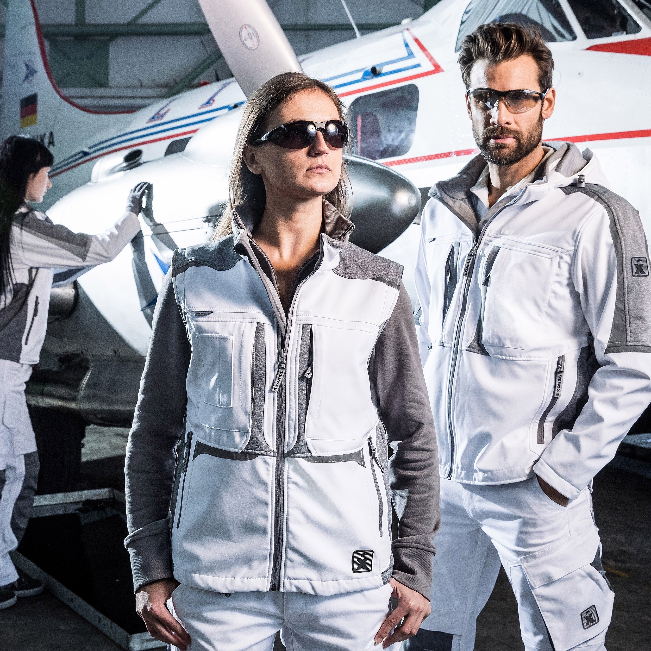 Weiße Arbeitskleidung mit grauen Details – moderne Berufsmode für Industrie & Handwerk, präsentiert vor Flugzeug in Werkstatt.