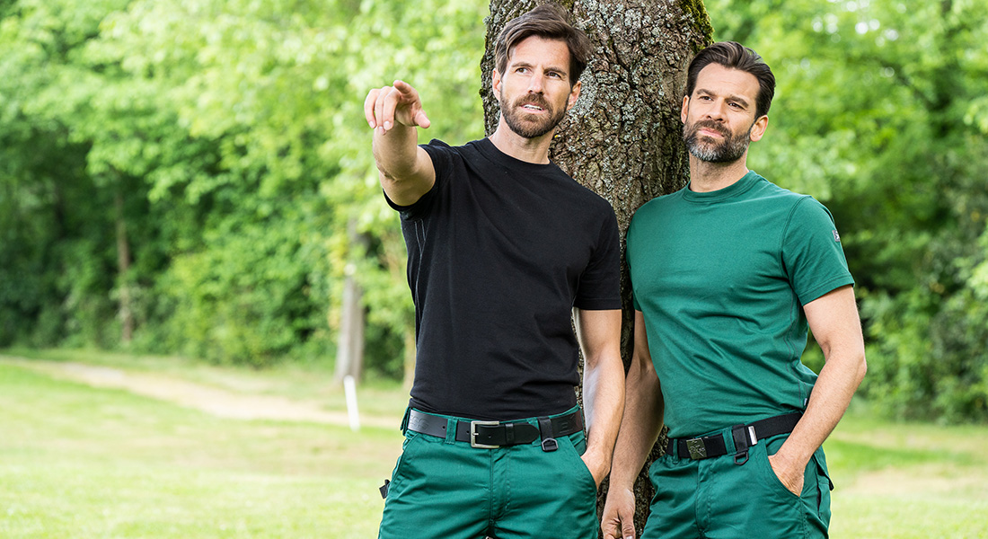 Zwei Männer in grüner Arbeitskleidung stehen im Freien vor einem Baum. Einer trägt ein schwarzes T-Shirt und zeigt in die Ferne, der andere lehnt entspannt am Baum.