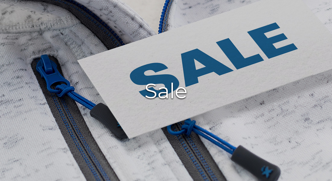 Detailaufnahme einer hellen Jacke mit blauen Reißverschlüssen, auf der eine weiße Karte mit der Aufschrift „SALE“ liegt. In der Bildmitte steht zusätzlich der Schriftzug „Sale“.
