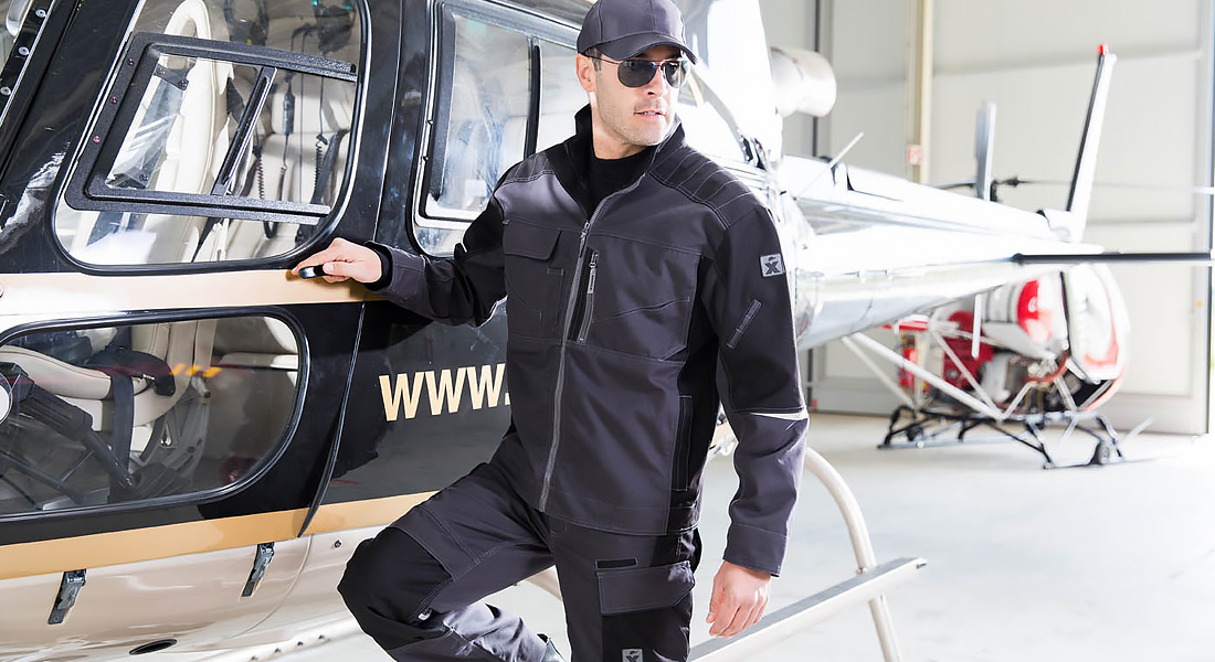 Mann in schwarzer Arbeitskleidung und Kappe steht neben einem schwarzen Hubschrauber in einer hellen Hangarhalle.