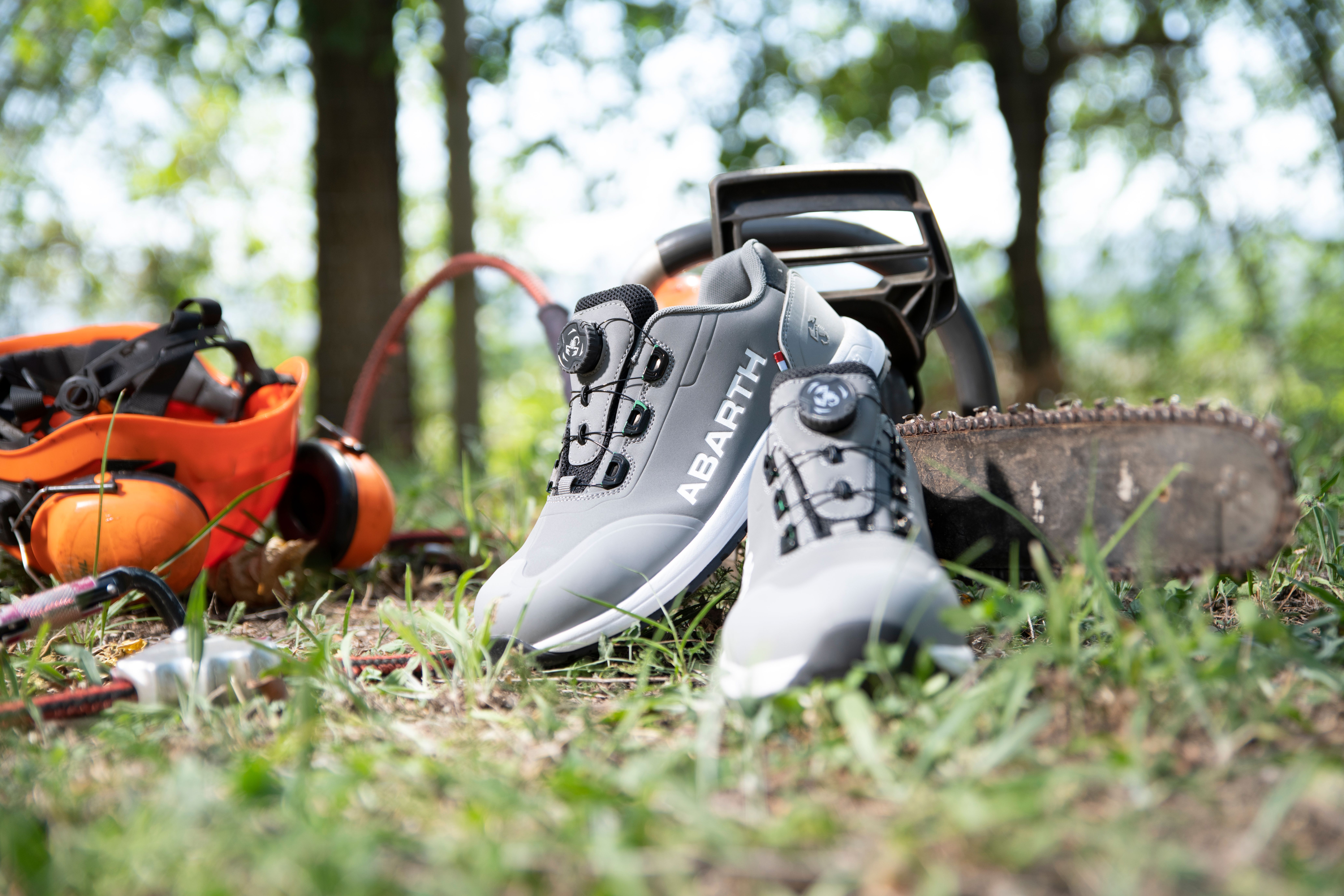 Graue ABARTH Sicherheitsschuhe mit schwarzer Sohle stehen im Gras neben Forstwerkzeug und Motorsäge.
