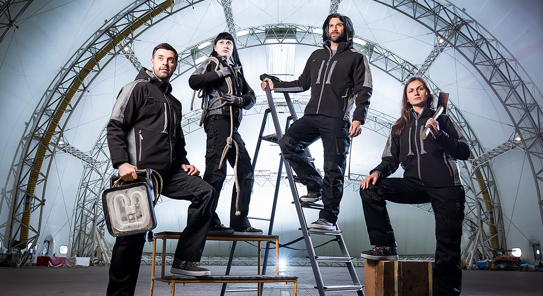 Vier Personen in schwarzer Arbeitskleidung posieren in einer großen Industriehalle. Zwei stehen auf Leitern, eine Person hält ein Werkzeug, eine andere eine Lampe.