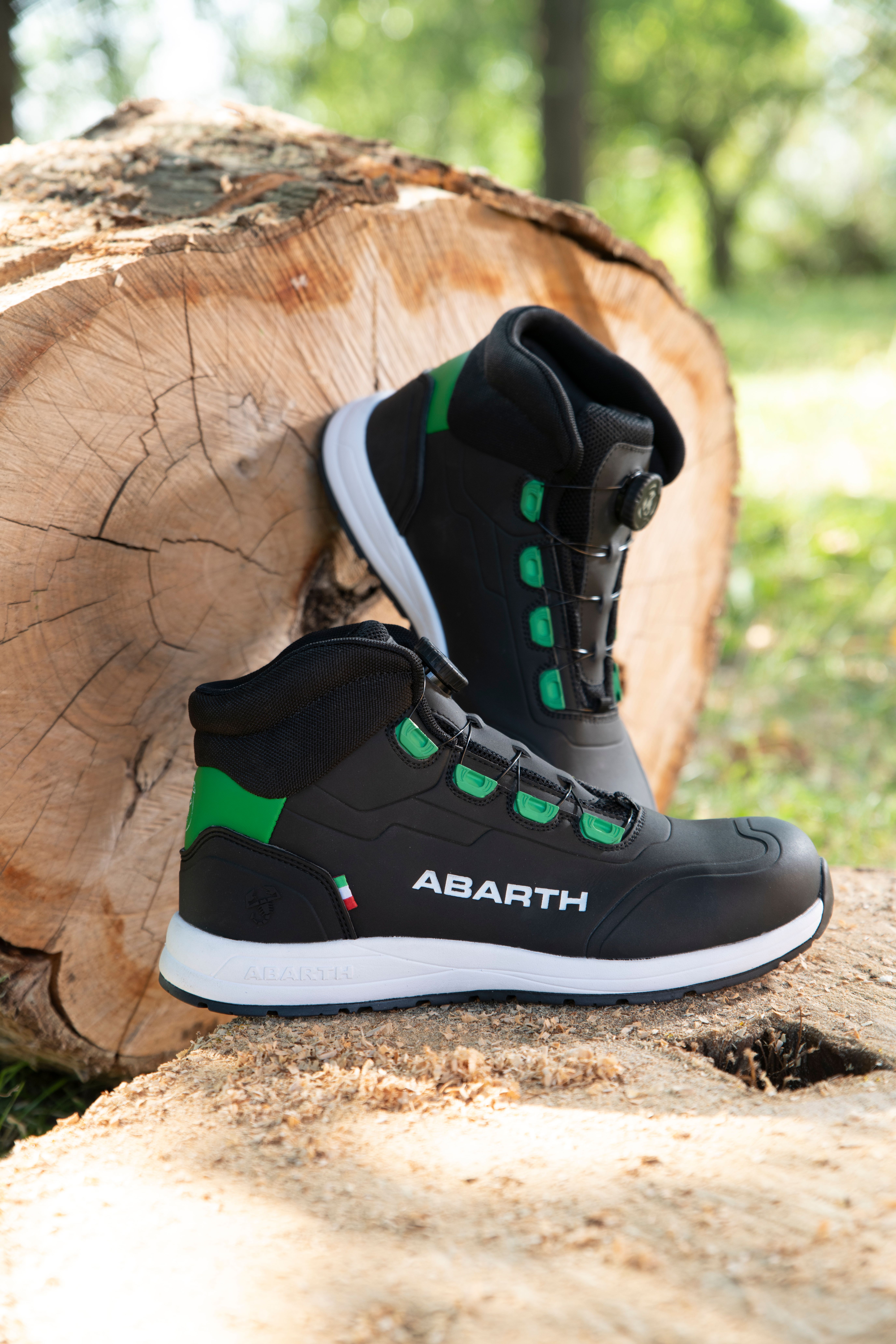 Schwarze ABARTH Sicherheitsschuhe mit grünen Details und weißer Sohle stehen auf einem Baumstamm im Freien.