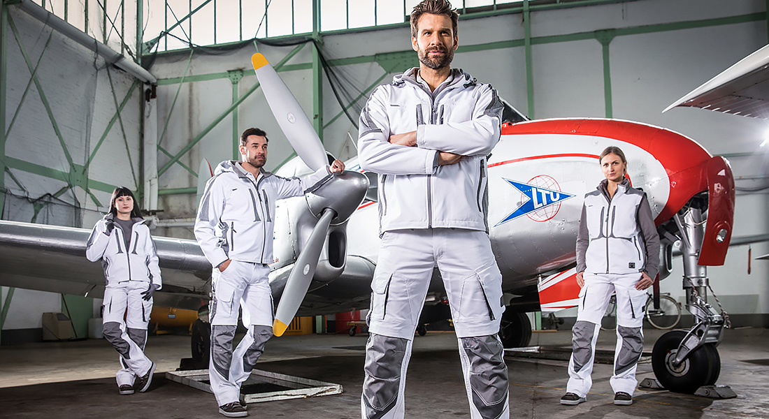 Vier Personen in weiß-grauer Arbeitskleidung stehen selbstbewusst in einem Hangar vor einem Flugzeug mit roter Spitze und Propeller.