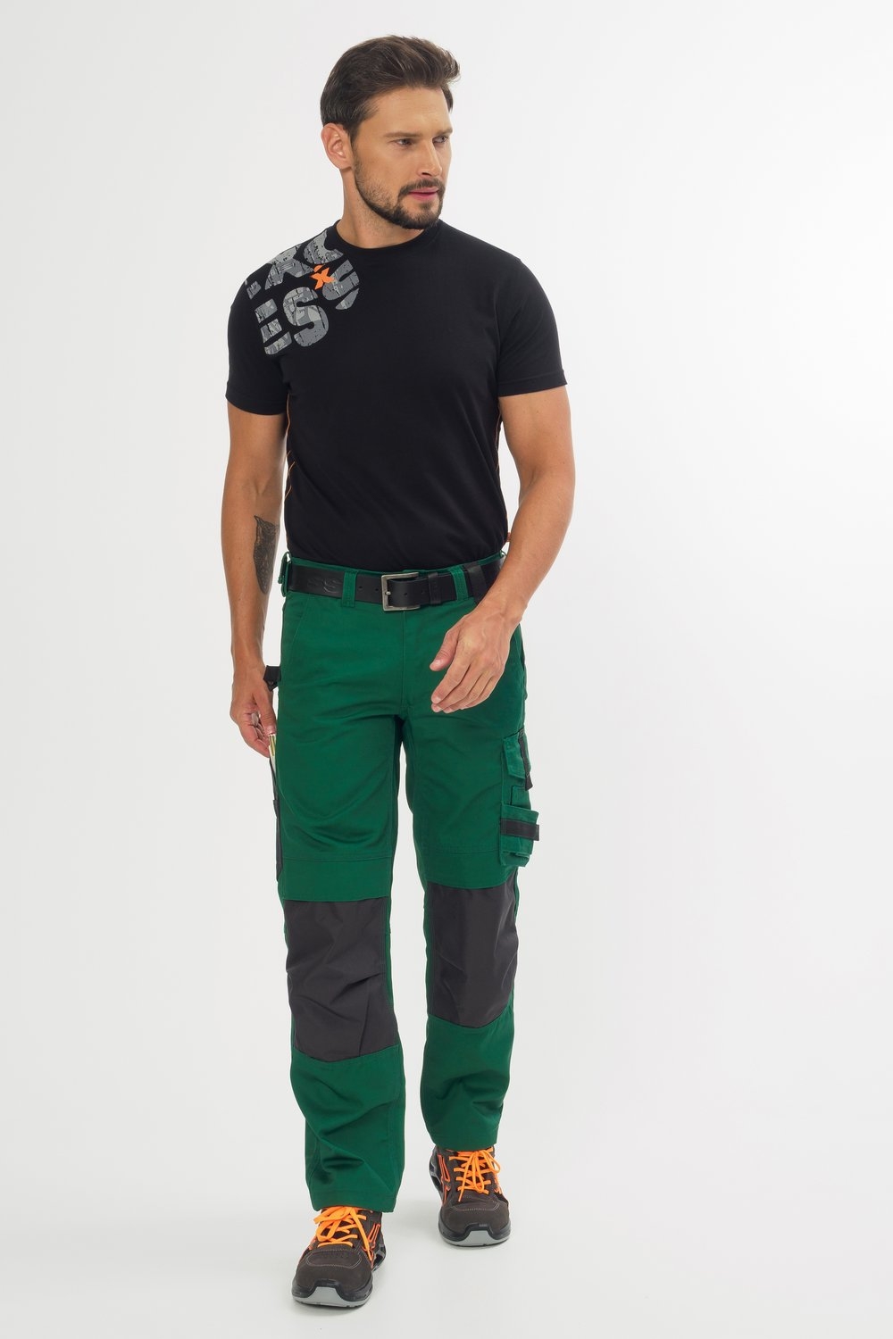 Mann trägt schwarzes T-Shirt und grüne Arbeitshose mit grauen Knieverstärkungen, dazu schwarze Arbeitsschuhe mit orangefarbenen Details.