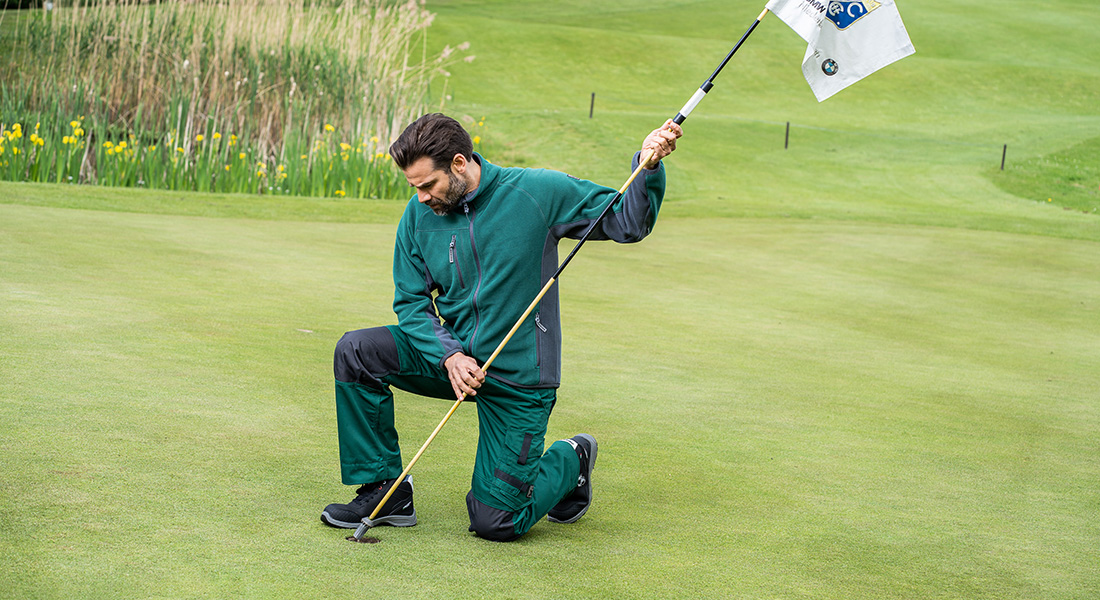 Mann in grüner Arbeitskleidung kniet auf einem Golfplatz und steckt eine Fahne in das Loch. Im Hintergrund ist gepflegter Rasen mit Schilfgras zu sehen.