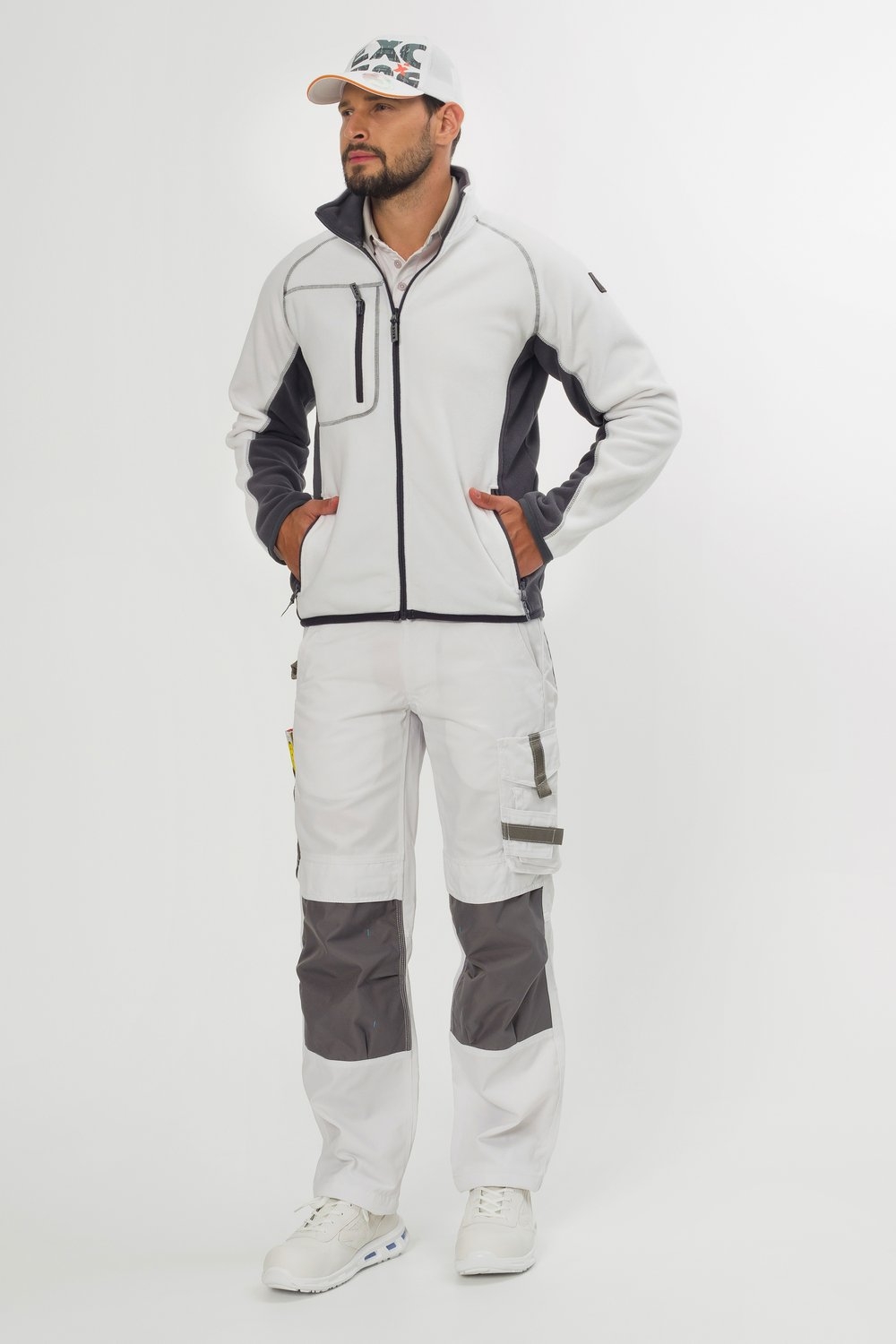 Ein Mann trägt weiße Arbeitsjacke mit Zippern und grauen Einsätzen, passende Arbeitshose mit Taschen sowie eine weiße Cap mit Logo.