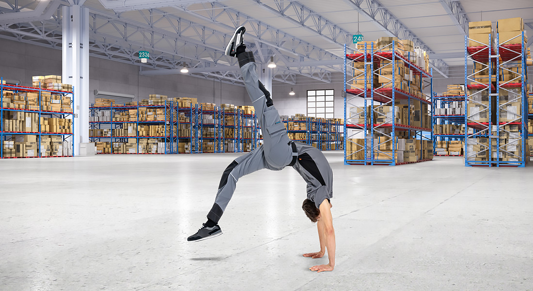 Mann in grauer Arbeitskleidung steht auf den Händen in einer großen Lagerhalle mit Regalen voller Kartons.