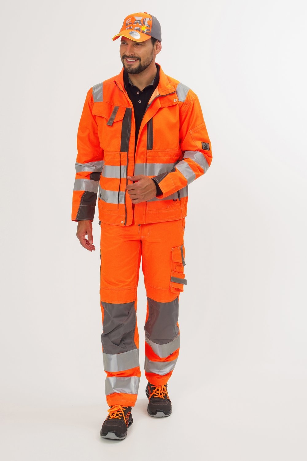 Mann in orangefarbener Arbeitsjacke und Hose mit silbernen Reflexstreifen, grauen Einsätzen und orange-schwarzen Sicherheitsschuhen.