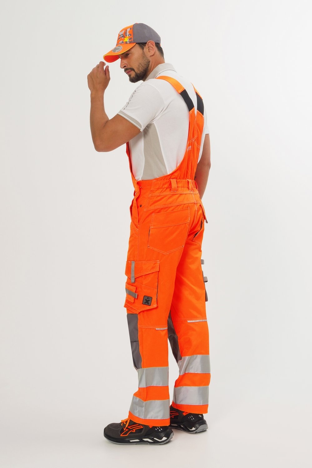 Mann von hinten in orangefarbener Latzhose mit silbernen Reflexstreifen, grauen Einsätzen, weißem Poloshirt und schwarzen Sicherheitsschuhen.