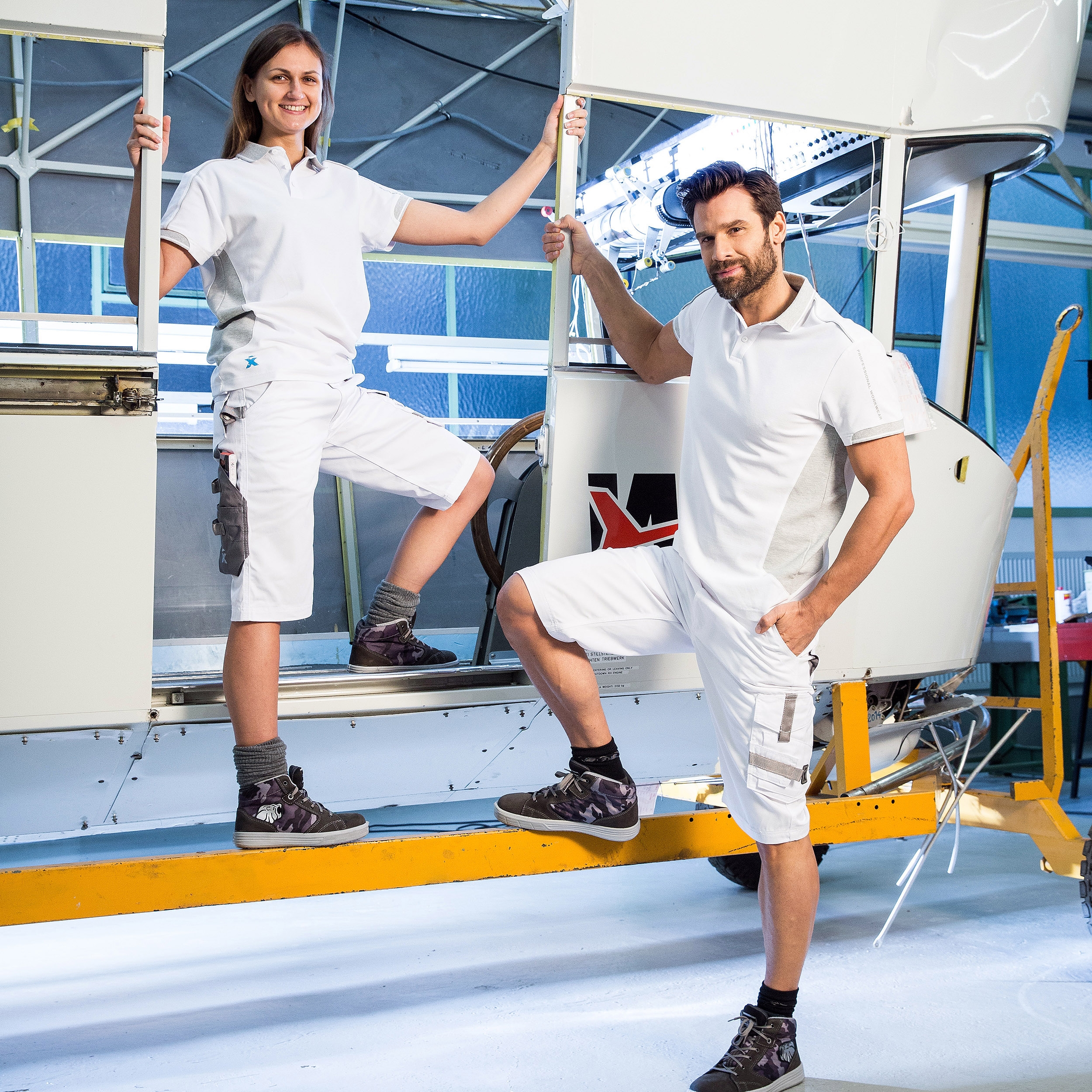 Arbeitskleidung weiß – Shorts und Poloshirt für Handwerk & Industrie, getragen von zwei Personen in Werkstattumgebung.