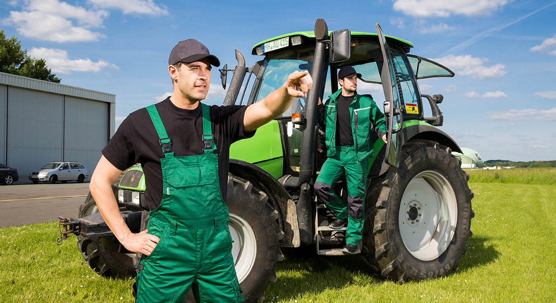 Zwei Männer in grüner Arbeitskleidung stehen bei sonnigem Wetter vor einem großen grünen Traktor auf einer Wiese. Einer zeigt mit ausgestrecktem Arm in die Ferne.