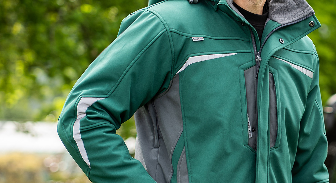 Ausschnitt einer Person in grüner Softshell-Arbeitsjacke mit grauen Einsätzen und reflektierenden Streifen, aufgenommen im Freien vor grünem Hintergrund.