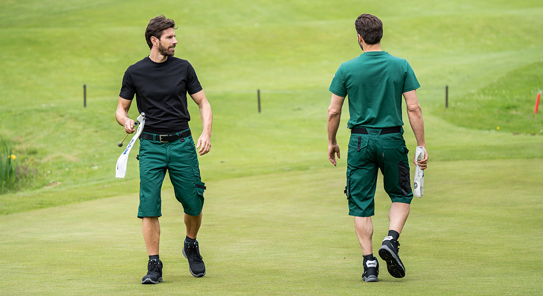 Zwei Männer in grüner Arbeitskleidung stehen auf einer gepflegten Rasenfläche, vermutlich einem Golf- oder Parkgelände. Einer schaut zurück, beide tragen Werkzeugtücher in der Hand.