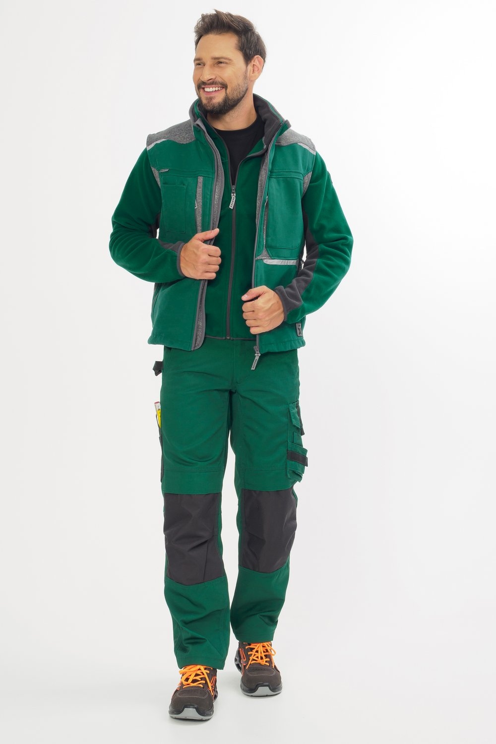 Mann trägt grüne Arbeitsjacke mit grauen Details und grüne Arbeitshose mit Knieverstärkungen, dazu Sicherheitsschuhe.