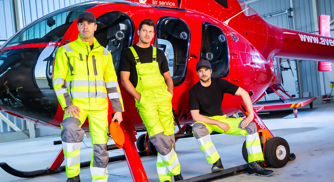 Drei Männer in leuchtend gelber Warnschutzkleidung stehen und sitzen vor einem roten Hubschrauber in einer Werkhalle.