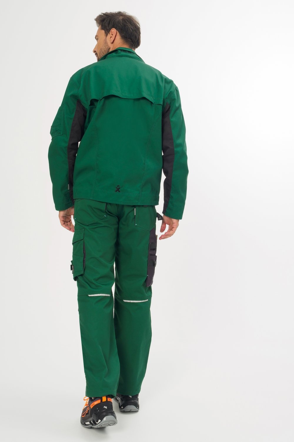 Mann in grüner Arbeitsjacke und Arbeitshose mit schwarzen Details, Rückansicht, Sicherheitsschuhe mit orangen Akzenten.