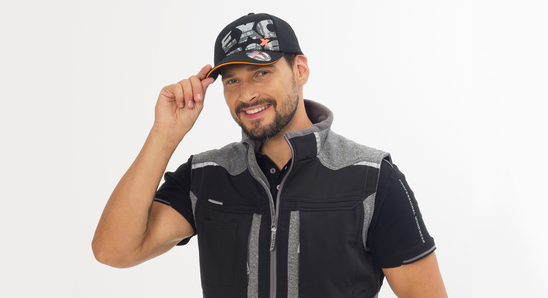 Mann mit Bart lächelt und tippt an den Schirm seiner schwarzen Kappe. Er trägt eine schwarze Arbeitsweste mit grauen Einsätzen vor hellem Hintergrund.