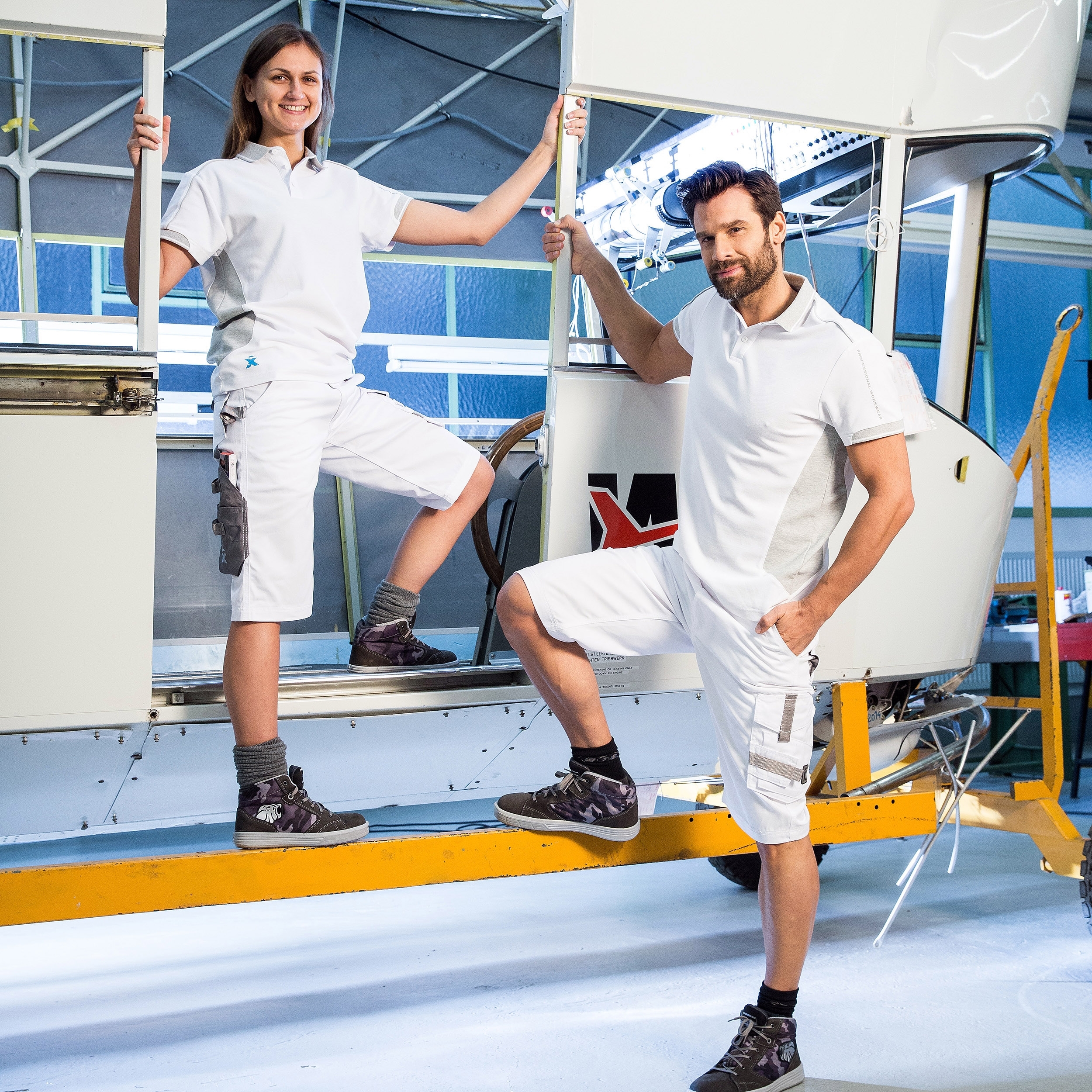 Arbeitskleidung weiß – Shorts und Poloshirt für Handwerk & Industrie, getragen von zwei Personen in Werkstattumgebung.