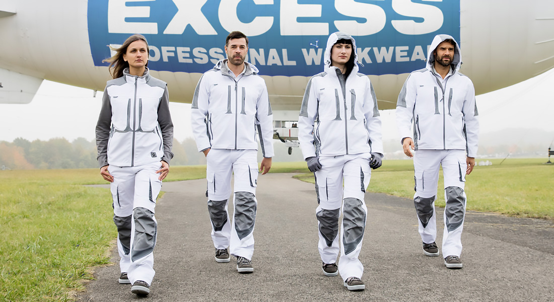 Vier Personen in weiß-grauer Arbeitskleidung gehen selbstbewusst auf einer Landebahn. Im Hintergrund schwebt ein Luftschiff mit dem Schriftzug „Excess Professional Workwear“.