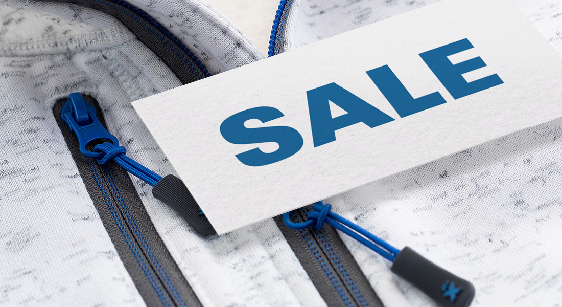 Nahaufnahme einer hellen Jacke mit blauen Reißverschlüssen, auf der eine weiße Karte mit der Aufschrift „SALE“ in blauer Schrift liegt.