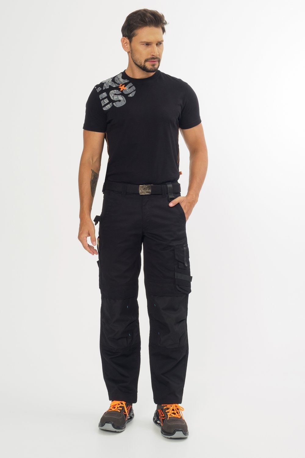 Ein Mann trägt schwarze Arbeitshose mit Seitentaschen und Gürtel, dazu schwarzes T-Shirt und schwarz-orangene Sicherheitsschuhe.