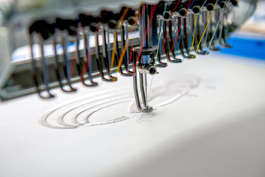 Nahaufnahme einer industriellen Stickmaschine, die ein gesticktes Motiv auf weißen Stoff aufbringt. Mehrere farbige Fäden sind in die Maschine eingespannt.