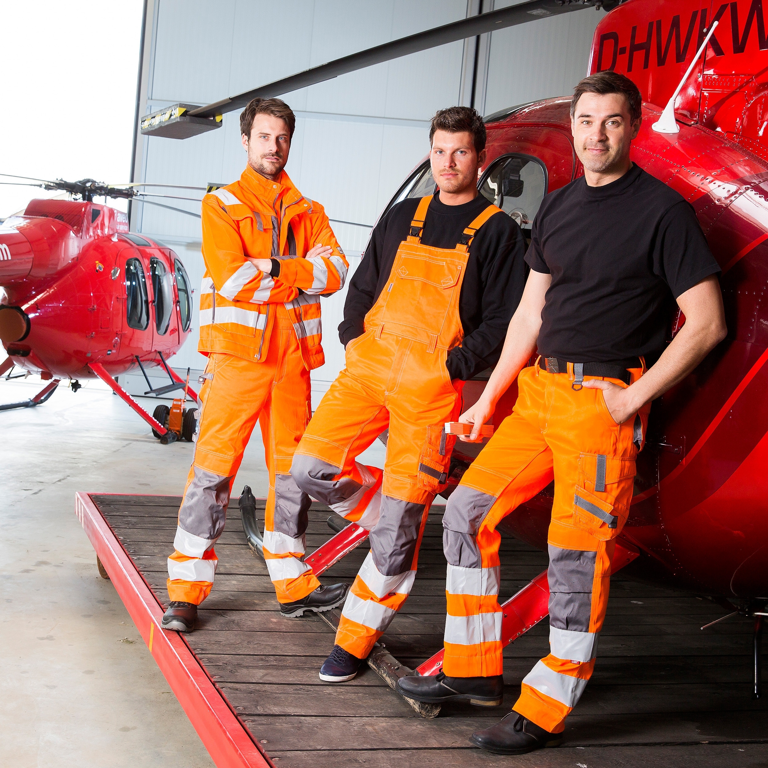 Gruppe von Männern in oranger Arbeits- und Warnschutzkleidung mit Reflexstreifen vor einem roten Rettungshubschrauber.